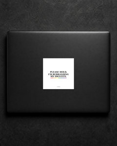 Please Hold, I'm Rebranding My Identity sticker centered on a black laptop lid.