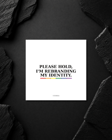 Please Hold, I'm Rebranding My Identity sticker on a dark studio surface.