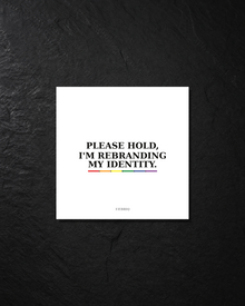 Close view of a Please Hold, I'm Rebranding My Identity sticker on a dark textured surface.
