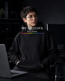 FÆBRIQ Off The Clock. Still Iconic. Heavyweight Hoodie — developer-workstation lifestyle view showing the white chest print and six-stripe Pride Circuit bar.